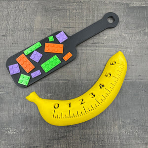 Een kleine BDSM-paddle in de kleur zwart met paarse, oranje en groene bouwsteentjes en een banaan