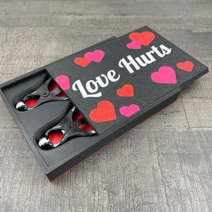 Een set zwarte tepelklemmen in een doosje met de tekst "Love Hurts"