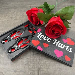 Een set zwarte tepelklemmen in een doosje met de tekst "Love Hurts" en twee rozen
