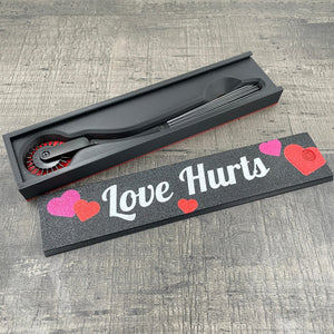 Een zwarte BDSM Wartenberg pinwheel in een doosje met de tekst "Love Hurts"