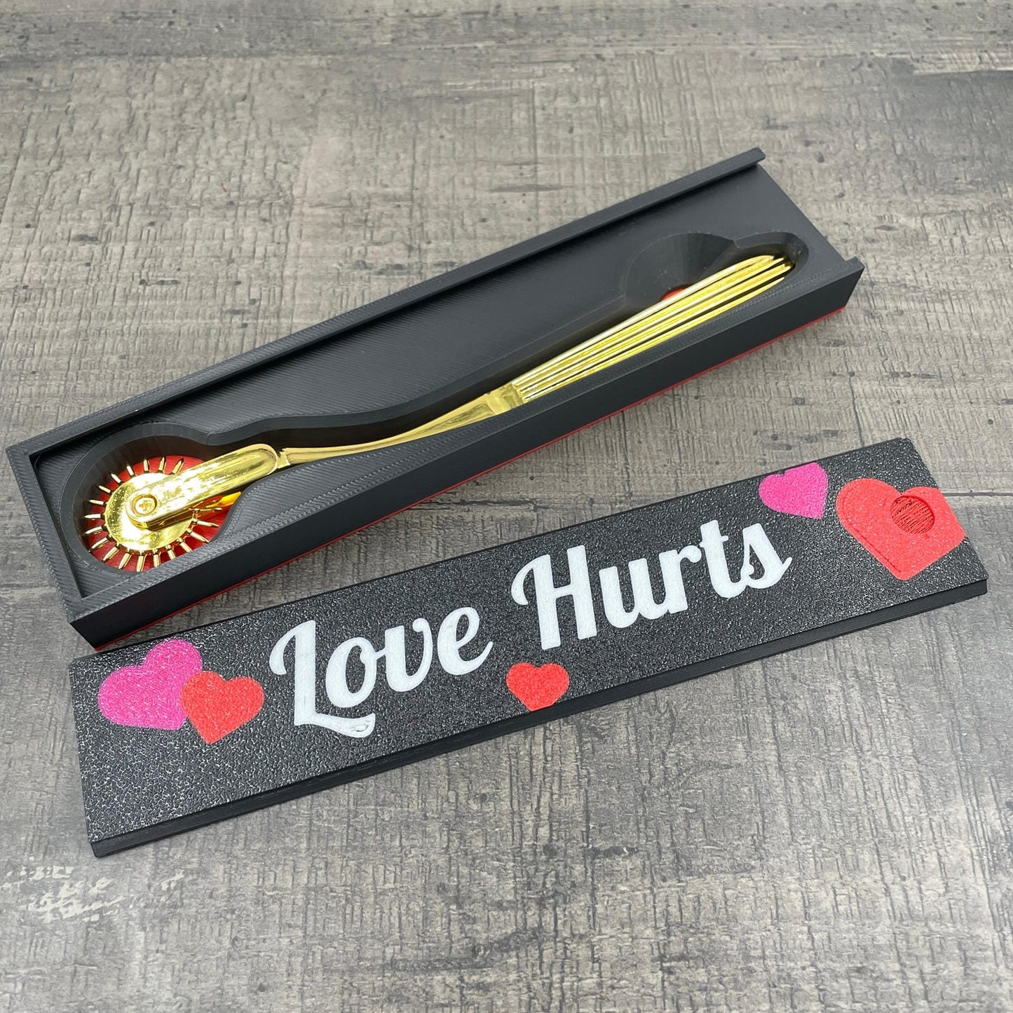 Een gouden BDSM Wartenberg pinwheel in een doosje met de tekst "Love Hurts"