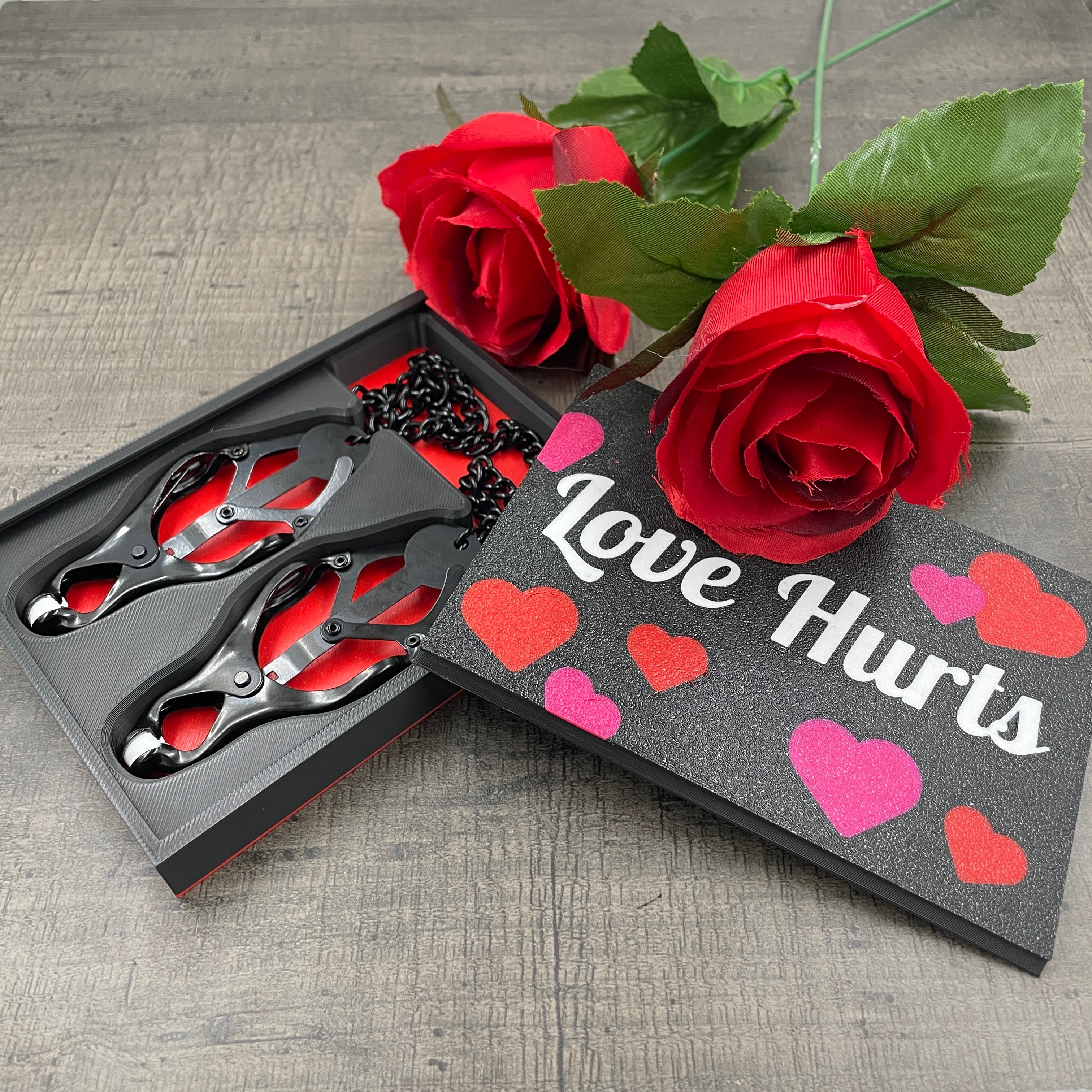 Een set zwarte tepelklemmen in een doosje met de tekst "Love Hurts" en twee rozen
