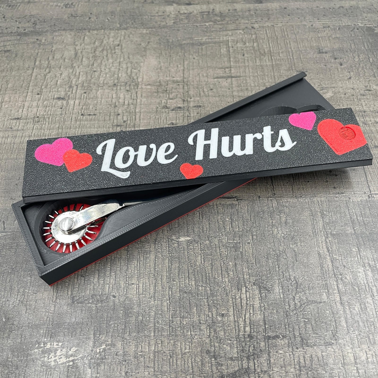 Een silverkleurige BDSM Wartenberg pinwheel in een doosje met de tekst "Love Hurts"