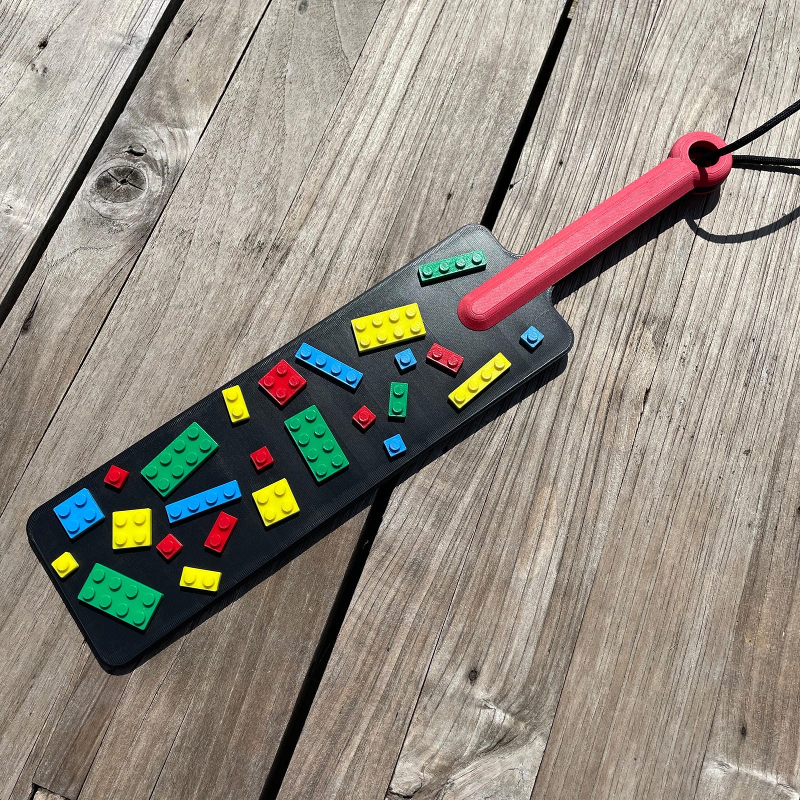Een BDSM paddle in zwart met gekleurde bouwsteentjes en een rood handvat